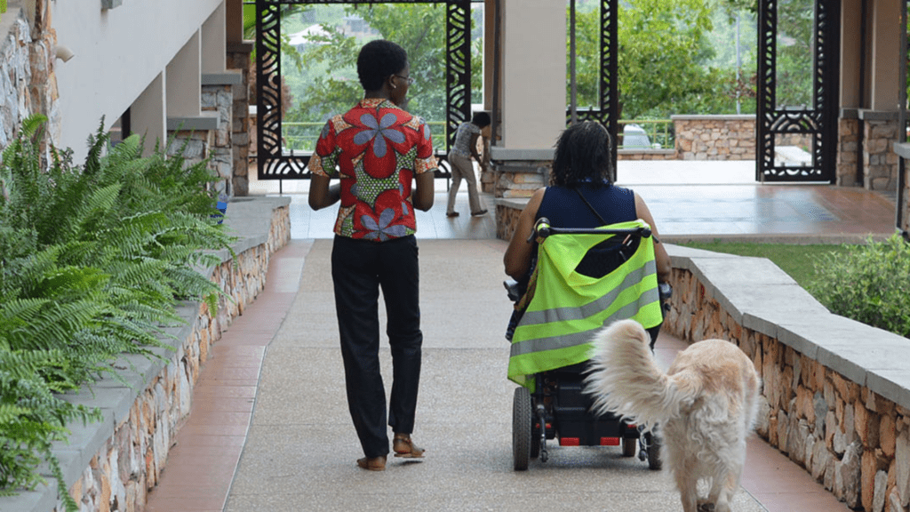 One man standing, one woman in a wheelchair and a dog next to them walking down a paved path.