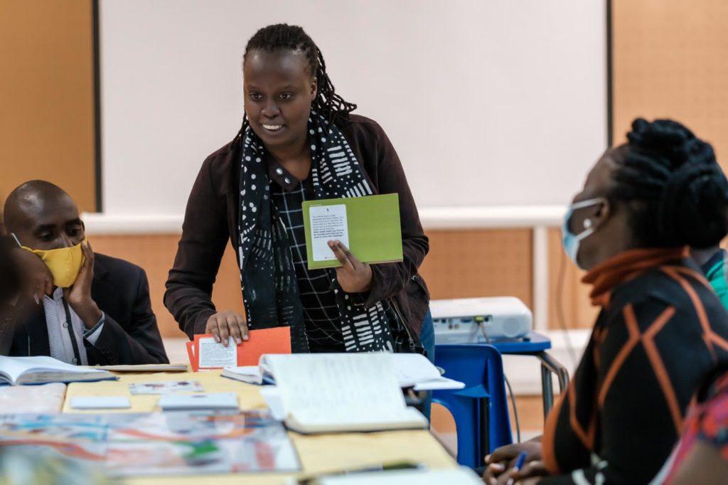 A facilitator is standing at the head of a table, holding a few cards in her hand