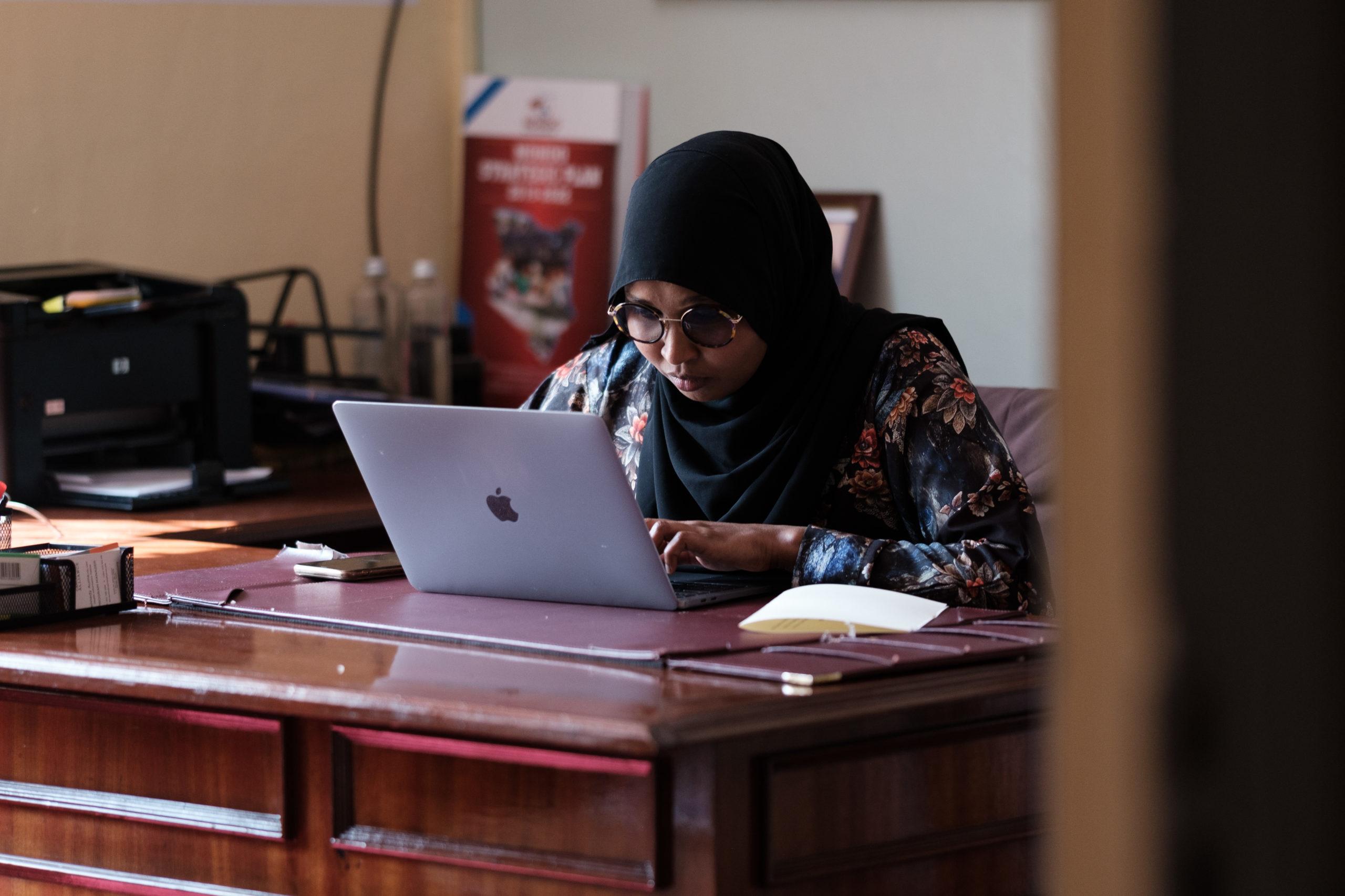 A picture of a woman wearing a hijab sitting at her desk and using her laptop