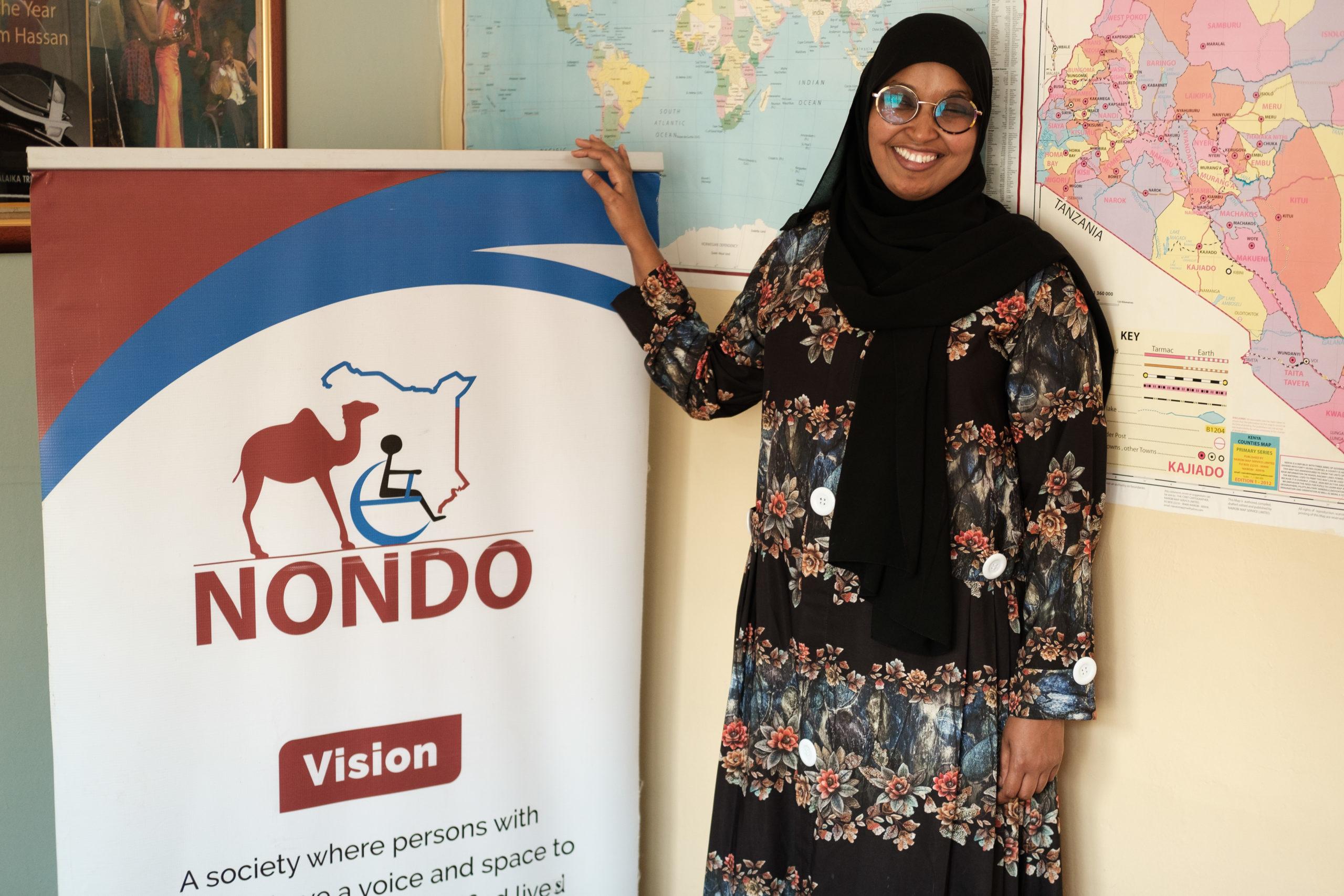 A picture of a woman wearing a hijab and glasses standing next to a banner with the logo for the organization she works at