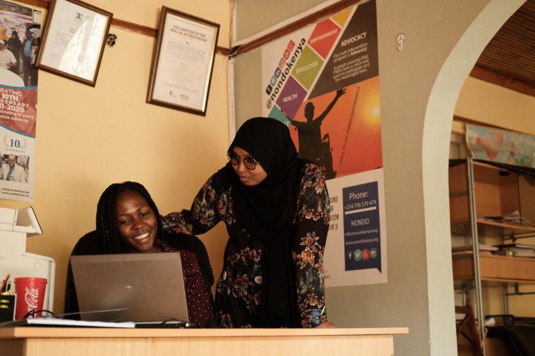 A photograph of two women in an office looking at a laptop and smiling. One woman is sitting and the other is standing looking over her sholder.