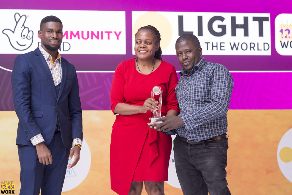 A woman in a red dress receives an award from a man in a blue suit onstage at an event. She is accompanied by a man in a blue shirt. The three of them smile and pose for the photo.