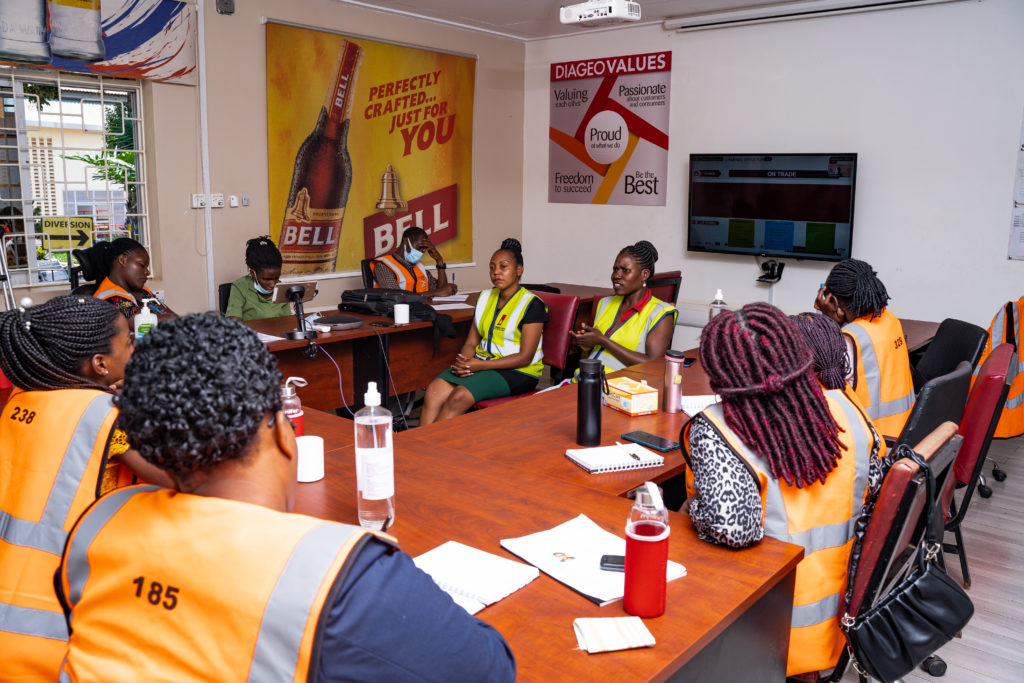 A group of people sitting around a table in the boardroom at Uganda breweries limited. All of the people are wearing orange safety vests. There is a poster for Bell Beer behind them.